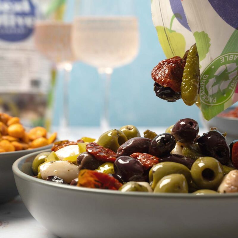 A 1kg bag of The Real Olive Company Antipasti olives being poured into a big party bowl. In the background you can see a bowl of Spicy Habas & Corn and two glasses of wine.